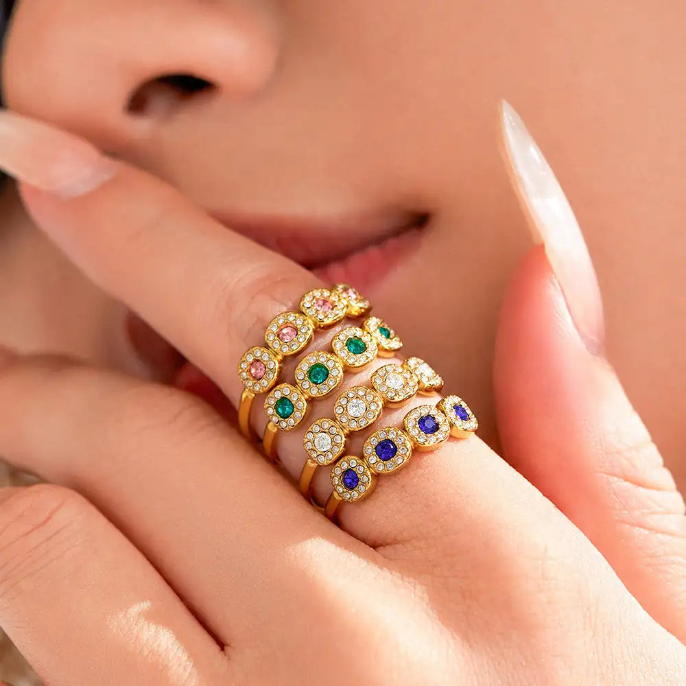 A set of five stacked gold rings, each featuring a central colored gemstone, pink, green, blue, and clear, and surrounded by smaller diamonds.